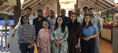 Ravi at a team event. A diverse group of about 12 people pose together, smiling. They appear to be in a restaurant with wooden beams and brick walls. The group includes men and women of various ages, wearing casual to semi-formal attire.