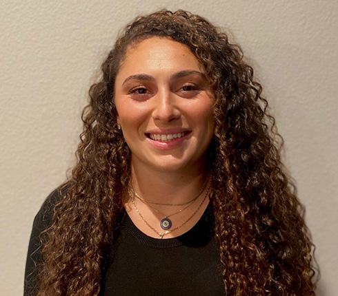 Raba, a female with long brown curly hair, is smiling at the camera wearing a plain black shirt.