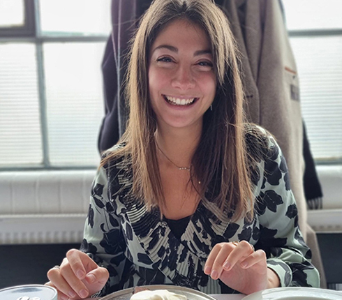 Rebecca, a female European, long brown hair, sitting in a restaurant with a dessert in front of her, she is smiling.