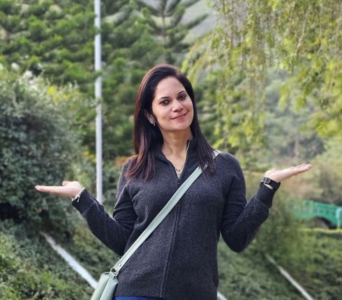 Ruchika stands in front of trees with her arms bent and palms facint upwards, she has dark hair and dark eyes and is smiling to the camer, she is wearing a black longsleeved top and has a light blue handbag across her upper body