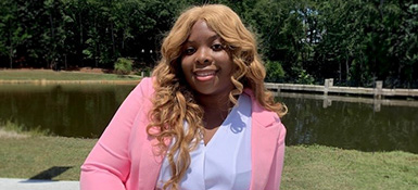 Ryan, a woman with blonde curly long hair, is sitting in front of a pond wearing a pink suit jacket and white button-down shirt