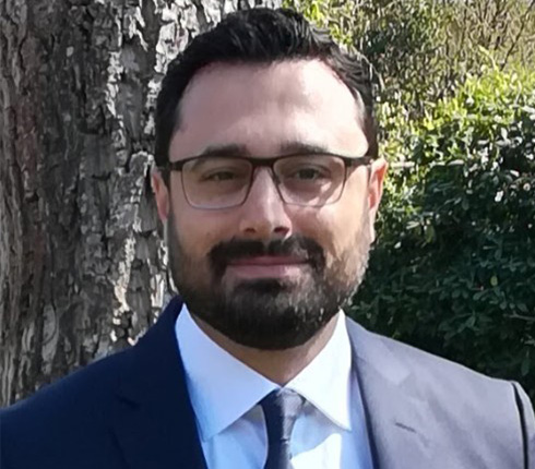 Man with brown hair, eyes, and a beard, wearing glasses looking directly in front; while wearing a black suit with a white shirt.