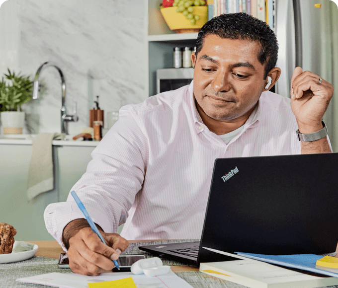 Imran, an Indian man HR professional at Parexel working from home at his kitchen table. He is in front of a laptop while making notes on paper next to it. He wears a pink striped shirt and has short dark hair.