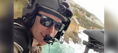 Thomas, an American man in a helicopter, wearing a helmet and sunglasses. You see a small part of the helicopter on the right side and out the window is a cliff landscape.