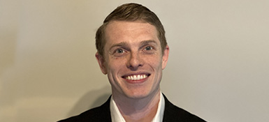 Thomas Geiger, IT Security Analyst, with blonde hair and smiling, wearinga white collared shirt with black jacket