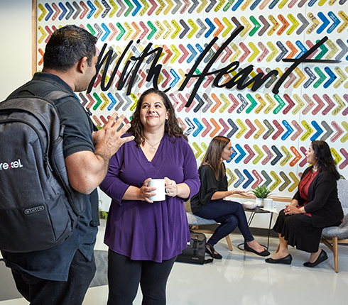 The two colleagues Imran and Cristina are in the front, two other colleagues are sitting in the back in front of a wall with a "With Heart" signage. Imran talks and Cristina reacts with a smile.