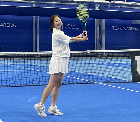 Yena, a female Korean, on a tennis court, wearing a white tennis outfit and holding up a tennis racquet.