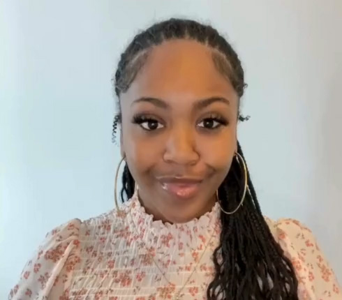 Alexis, a Black-American woman with braided hair and hoop earrings, wearing a white and pink floral top with a high neckline. She has a friendly expression against a light background.