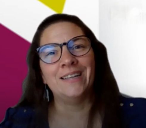 Jennifer speaking to the camera. She has long brown wavy hair, wearing glasses and dangly earring is smiling at the camera.