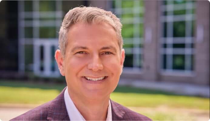 Headshot of Chris Learn, a middle-age white male, with short grey hair, he is smiling at the camera and he wears a suit. In the background is a building and some grass.