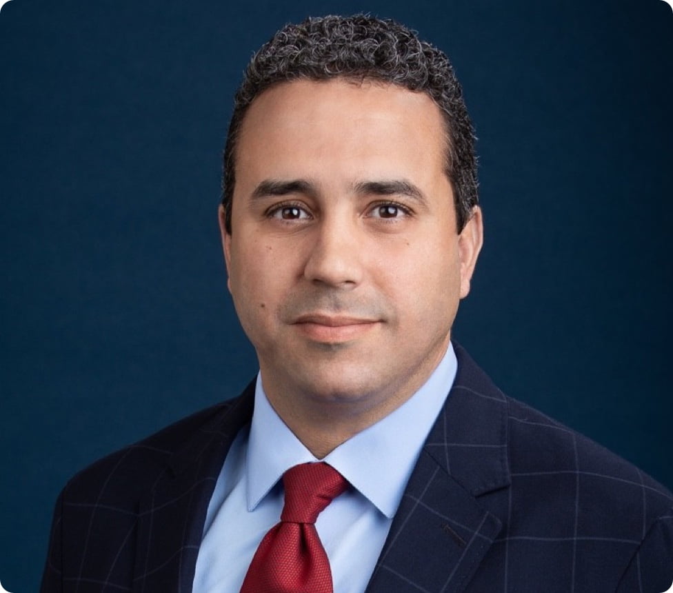 Professional headshot of Hicham, he is a middle-aged male with Arabian descent, dark, curly, short hair, her wears a suit with a tie.