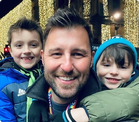 Ben, a male white British and Senior Regulatory Affairs Consultant at Parexel, with his two children, one is hanging onto his one shoulder, the other is looking over his other shoulder, in the background are festive lights, they all look at the camera.