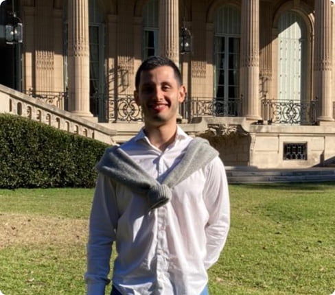 Picture of Daniel, Project Leader at Parexel, is standing in front of an old building with columns, he is smiling at the camera, he has short hair and the sun is coming from the side, he is wearing a white shirt
