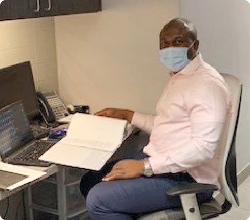 Picture of Emmanuel, Senior Clinical Research Associate at Parexel, sitting at a desk while visiting a site, documents and a computer in front of him, he is wearing a mask, and is looking at the camera.