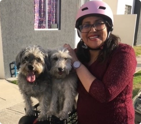 Picture of Itzel, Senior Clinical Data Analyst, standing next to her two dogs, holding them while they are on the back of her bike, she is wearing a pink bike helmet and smiles into the camera.