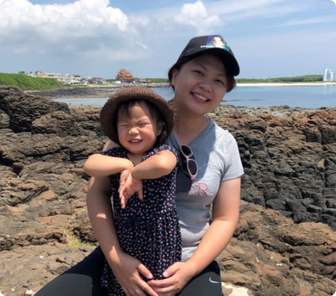 Joy, a female Taiwanese and Senior Director, Statistical Programming at Parexel, she is kneeling behind her child having her arms around her, they are outside on rocks in front of water, she is smiling at the camera, and the child is making a joyful face.