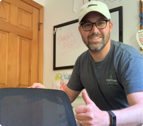 Judson, a male white American and Global Category Sourcing Manager, standing behind his desk chair he received from Parexel, smiling and holding a thumb up, in the background is a white board with the words "Thanks FWA!!" (Flexible Work Arrangement)