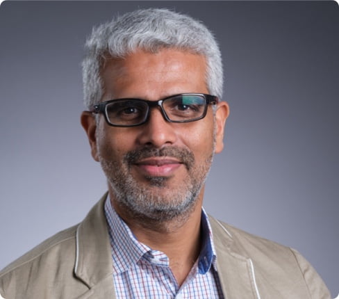 Picture of Sanjay, Executive Vice President, Safety & Logistics and Country Head of Parexel India, it's a classic headshot in front of a grey background, he is wearing glasses, a shirt, and a beige jacket.