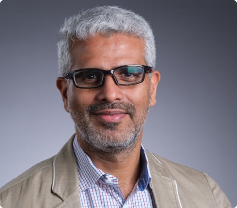 Sanjay, a male Indian and Executive Vice President, Safety & Logistics and Country Head of Parexel India, it's a classic headshot in front of a grey background, he is wearing glasses, a shirt, and a beige jacket.