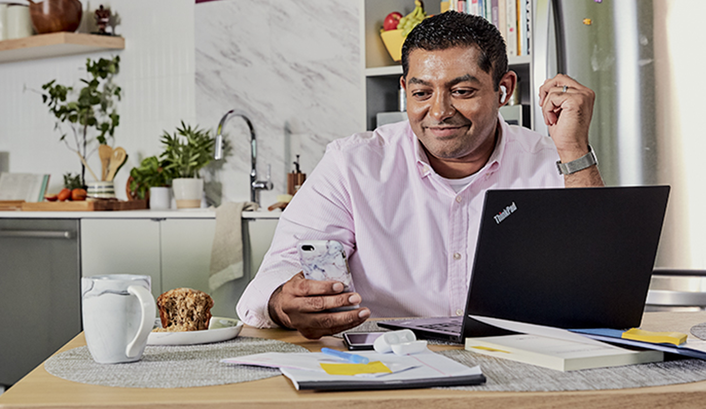 Middle-age, Indian male working from home, he is looking at his phone, laptop is also in front of him, in the background is a kitchen.