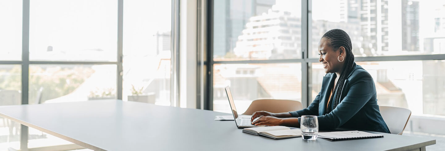A woman in a business suit is sitting at a large desk, and typing on a laptop while smiling.